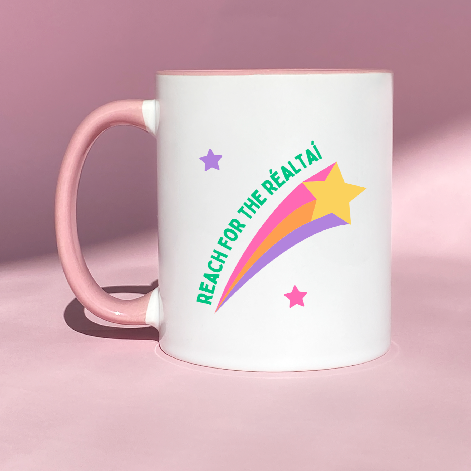 White ceramic mug with a colorful shooting star design and scattered stars, featuring the inspiring Irish phrase 'Reach for the Réaltaí.